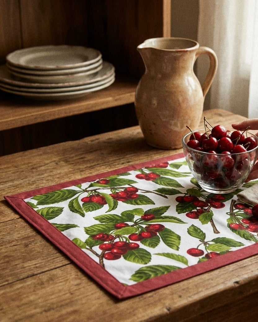 Person holding a bowl of cherries on a wooden table with a cherry-themed placemat.