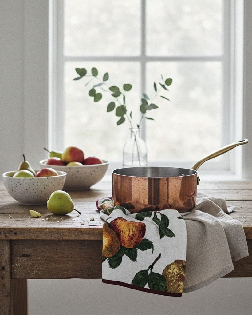 Copper pot on a wooden table with fruit and a floral towel in a bright room.