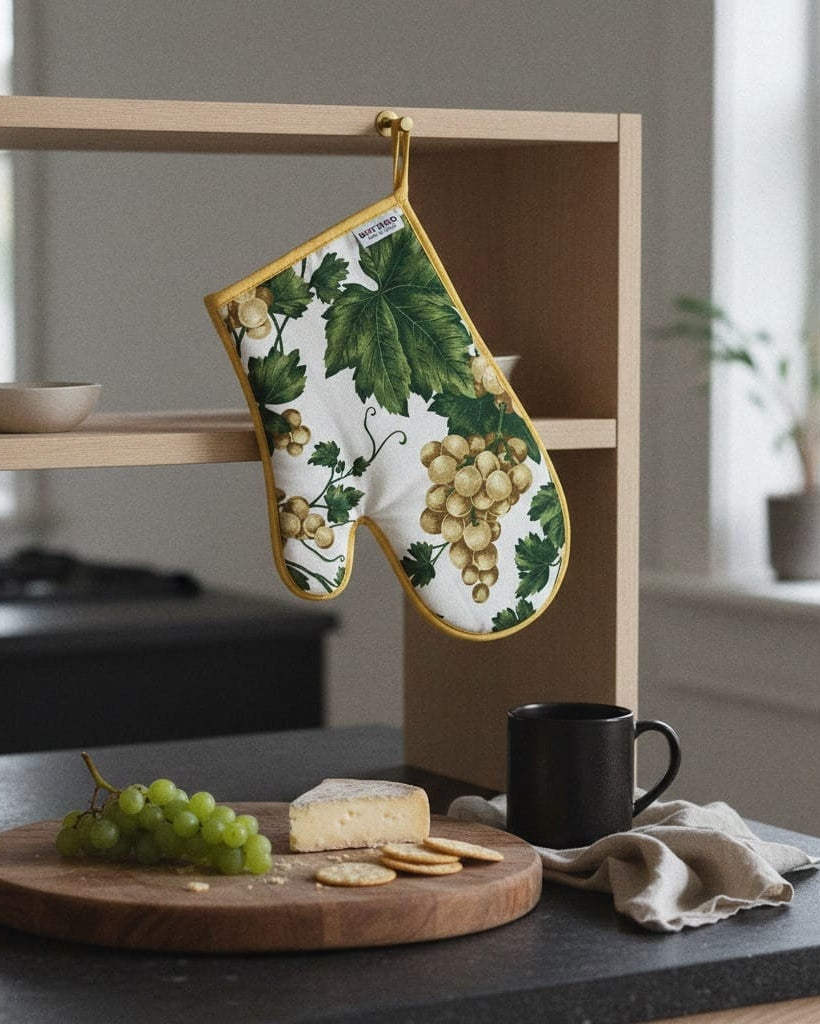 Oven mitt with grape design hanging on a kitchen shelf with a cutting board and snacks below.