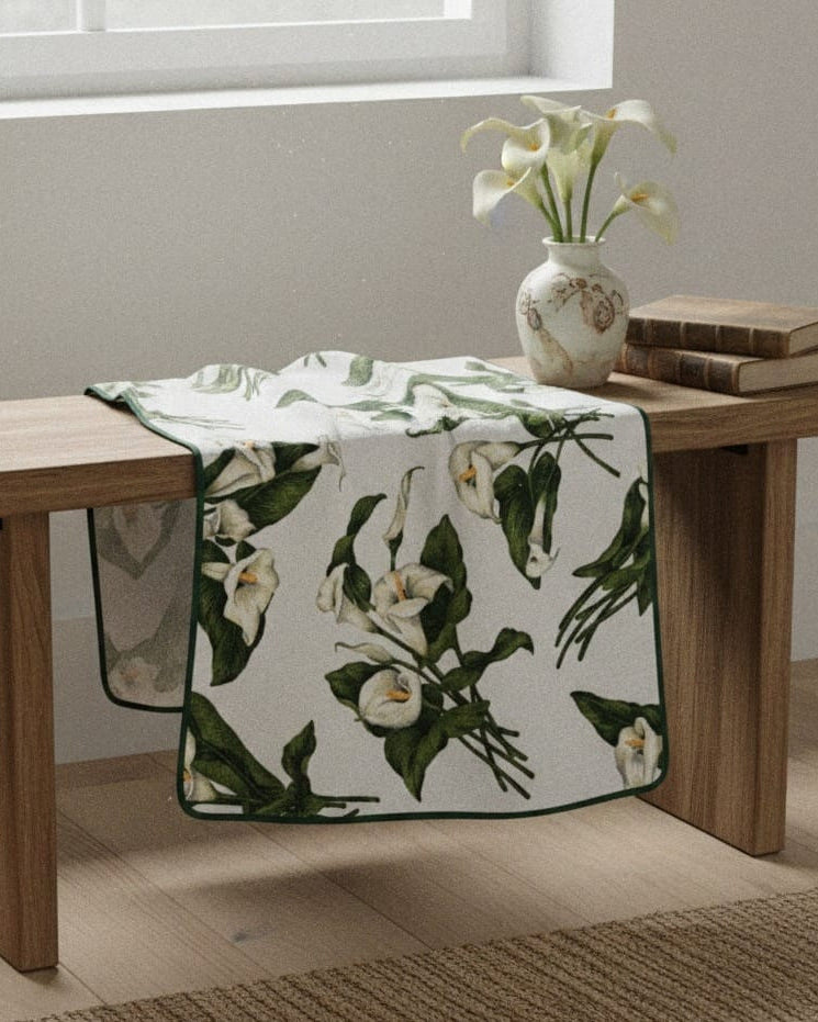 Floral-patterned blanket draped over a wooden bench with a vase and books on a light-colored floor.