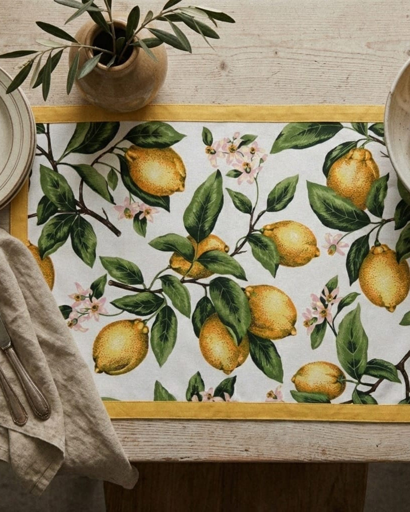 Table setting with a lemon-patterned placemat, bowl of lemons, and potted plant on a wooden table.